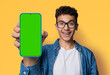 Shocked happy man wear glasses braces, open mouth, show cell phone, smartphone, mobile phone, cellphone green chroma key mockup mock screen, isolated yellow wall background. Online offer ad.