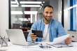 © Tetiana - Portrait of a smiling young Hispanic man sitting in a modern office at a desk, working with documents, using a laptop and a calculator
