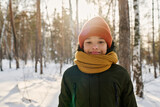Fototapeta  - Adorable boy in knitted yellow scarf and brown beanie hat looking at camera with smile while standing in natural environment