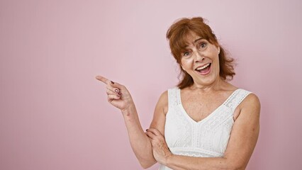 Wall Mural - Cheerful middle age woman smiling wide and pointing to the side, confidently showcasing something off camera. dressed to impress, standing against a vibrant pink backdrop.