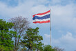 Thailand flag waving in the wind over green trees with blue sky background.