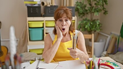 Poster - Middle-aged artist woman in studio, holding paintbrushes, amazed and scared with open mouth in surprised disbelief