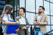 © Srdjan - Three young business people talking to each other while standing outdoors of office building with suitcase, laptop and register. Group of professional workers discussing about job and plans outside