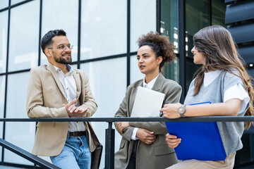 Wall Mural - Three young business people talking to each other while standing outdoors of office building with suitcase, laptop and register. Group of professional workers discussing about job and plans outside