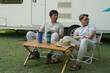 Two Asian male friends carry camping picnics in their RV and sit and relax, chatting in the forest.