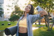 © Strelciuc - Adult woman holding yoga mat and using smartphone after workout in the park. Portrait of smiling female in sportswear walk using smartphone. Taking selfie.