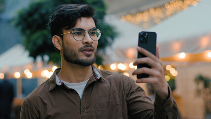 Handsome successful Indian Arabian ethnic man male student gen z guy businessman vlogger influencer talking recording online video call using gadget mobile cell phone app walking evening city street