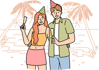 Honeymoon happy couple drinking champagne, standing on beach of sunny resort, wearing birthday caps. Romantic guy and girl celebrate beginning of honeymoon on ocean shore with palm trees