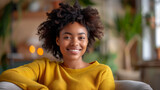 Fototapeta  - A woman with curly hair is sitting in a chair and smiling. She is wearing a yellow shirt. a young African American woman sitting in her chair, exuding happiness as she smiles warmly.