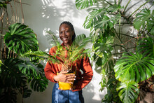 Happy African American Woman Plant Lover Holding Houseplant Cycas In Yellow Pot Smiling Looks At Camera In Sunny Day. Portrait Of Joyful Successful Black Female Owner Of Flower Shop Holding Houseplant