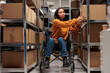 Woman wheelchair user taking cardboard box from shelf to prepare customer order for dispatching in warehouse. Asian package handler holding parcel, working in disability friendly storehouse