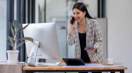 Wall Mural - Asian woman entrepreneur busy with her work in the office. Young Asian woman talking over phone or cellphone while sitting at her desk.
