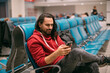 A young, handsome man is sitting with a phone in his hands at the airport in the evening. A guy, a tourist with a smartphone and a backpack is waiting for departure at the gate