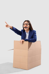 Poster - Young businesswoman with funny disguise sitting in cardboard box and pointing at something on grey background