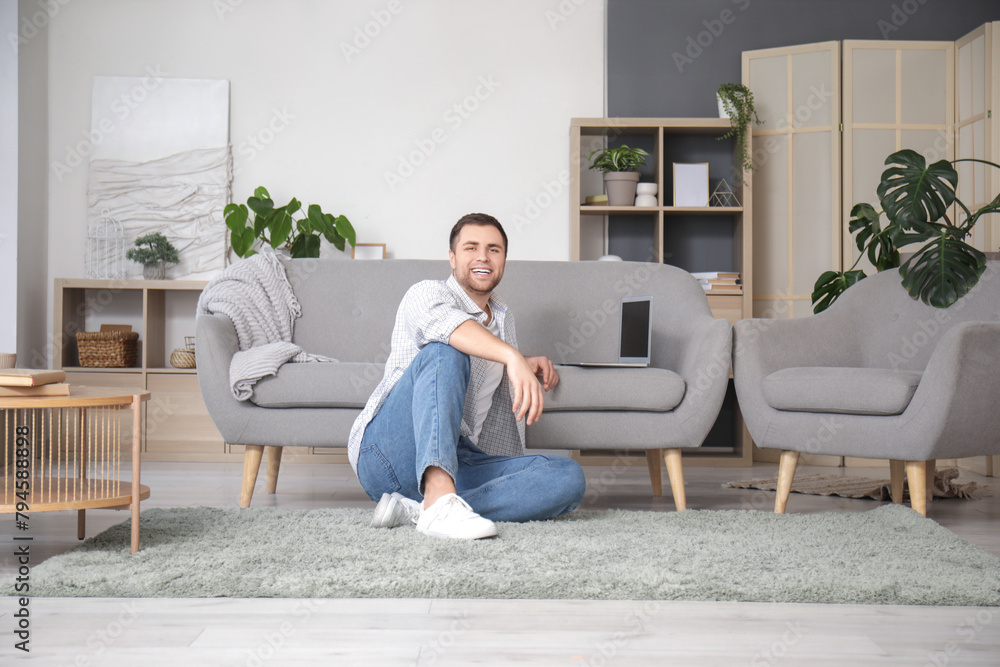 Young man sitting beside grey sofa with laptop at home