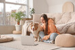 © Pixel-Shot - Young woman with cute fluffy Australian Shepherd dog and laptop sitting near sofa in living room
