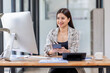 © David - Young happy professional business asian woman employee sitting at desk working on laptop in modern corporate office interior. Smiling female worker using computer technology typing browsing web.