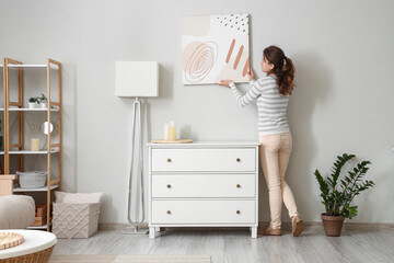 Wall Mural - Young woman hanging picture near chest of drawers in stylish room