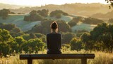 Fototapeta  - A woman sits on a bench looking out at the rolling hills and distant trees back turned to the camera as practices meditation . .