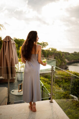 Back view Young beautiful woman enjoying a healthy raw fruit vegetable juice while relax at hotel resort by the sea, view from ocean cliff