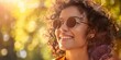 Smiling young woman with curly hair wearing round sunglasses basks in golden sunlight.