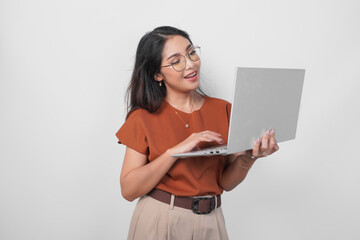 Sticker - Smiling young woman wearing brown shirt and eyeglasses while holding laptop to work or browsing isolated over white background.