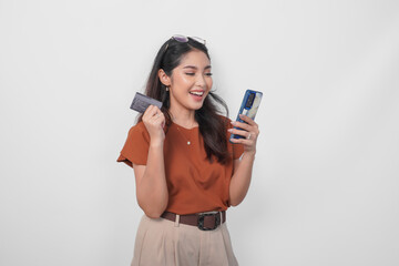 Wall Mural - Beautiful young Asian woman wearing brown shirt is smiling while holding her smartphone and credit card over isolated white background.