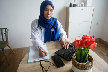 Serious Asian muslim woman doctor using laptop and writing notes in medical journal, sitting at desk