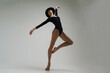 young ballerina in a black bodysuit shows ballet steps in motion standing on one leg and spreading her hands in a hat