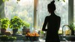 A woman standing in a kitchen looking out the window. There are plants and fruit on the counter. The light from the window is casting shadows over the woman and the plants.