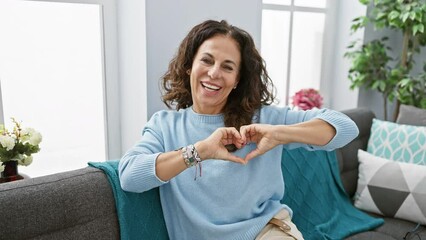 Poster - Heart-warming portrait of a middle-aged hispanic woman, sitting on a living room sofa, making a hand heart symbol. a romantic concept, showcasing a joyful surprise for valentine's at home.