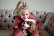 Beautiful blond girl child sits at home on a sofa, playing with and hugging dog, creating heartwarming moments of joy and companionship with pet