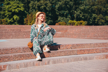 Young beautiful smiling blond hipster woman in trendy summer hippie costume clothes. Carefree female posing in the street. Positive model outdoors at sunset. Cheerful and happy in hat. sits at stairs