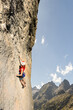 © VICTOR - A woman is climbing a rock wall with a red shirt and white helmet. The mountain in the background is covered in trees