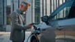 © inthasone - A man in a suit is charging an electric car. A man with short beard is standing near the modern silver SUV. He holds a mobile phone in his hand, outside an office building background.