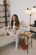 © polinaloves - Young woman using her smartphone and drinking tea, relaxing in a cozy living room.