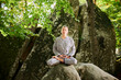 Woman practicing yoga outdoors in forest. Barefoot female on yoga mat surrounded by trees and large rocks, which suggests peaceful, natural environment ideal for meditation or yoga practice.