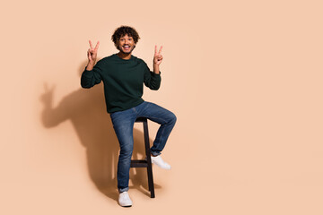 Poster - Full length photo of positive guy dressed sweatshirt sitting bar chair showing v-signs emtpy space isolated beige color background