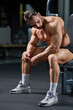 Serious Caucasian bodybuilder having rest after exercising in gym. Side view of tired male athlete with strong body sitting on chair, leaning on legs, looking down. Concept of bodybuilding, emotions. 