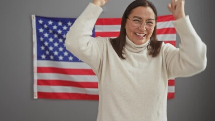 Canvas Print - Excited middle-aged hispanic woman, wearing glasses, bursting with victorious joy in the office, standing proud and screaming, raising arm in triumph, celebrating success near usa flag