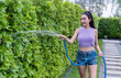Amidst the lush surroundings of her garden, a woman smiles joyfully as she waters a dense green hedge using a blue garden hose, enjoying the simple pleasures of gardening. Minimal free time leisure.