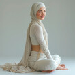 A Muslim arab woman dressed in a white hijab sits in the lotus position against a gray background. Square image
