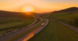AERIAL, LENS FLARE: Meandering motorway glows in orange hue of setting summer sun. Highway with smooth flowing traffic winds past green fields and lush forests stretching across the hilly countryside.