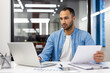© Tetiana - Concentrated and serious Arab young man sitting in the office and working with data, typing on a laptop and holding documents in his hands