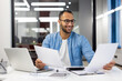 © Tetiana - A successful and smiling young Muslim man in glasses is sitting in the office at a desk with a laptop and working with documents and papers
