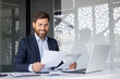© Tetiana - Portrait of a smiling young businessman sitting at an office desk with a laptop and looking through documents and bills