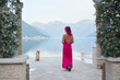 A woman with pink hair in a flowing dress stands facing a breathtaking lakeside view, with a Shiba Inu looking towards her