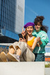 Vertical Screen: Two Young Women Share A Moment Of Laughter, Engaging With Smartphone Outdoors, Looking at Camera And Showing Peace Sign. They Are Dressed In Vibrant, Casual Attire. Friendship Concept