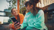 Two Young Women Sharing A Joyful Moment Outdoors, One Holding A Smartphone, Both Dressed In Vibrant Casual Wear, Embodying Friendship And The Digital Age Connection Amidst An Urban Setting.