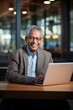 © jureephorn - Smiling senior Indian business executive sitting at desk using laptop.